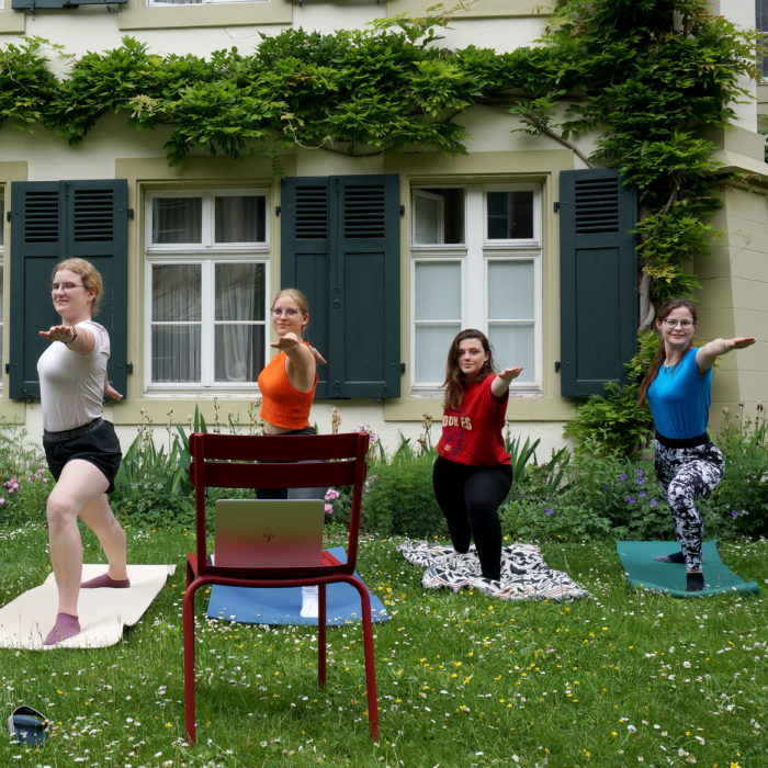 Vier Frauen machen auf dem Rasen Yoga, vor ihnen ein Stuhl mit einem Laptop.