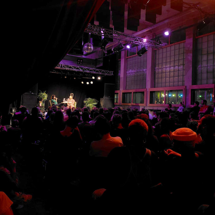 Im Vordergrund Publikum, im Hintergrund zwei Personen auf beleuchteter Bühne.