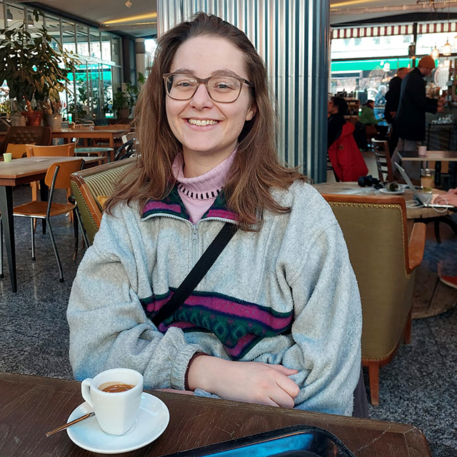 Junge Frau lächelt in Kamera, sitzt in Café mit Kaffeetasse vor ihr.